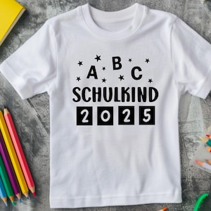 May include: White t-shirt with the text "A B C SCHULKIND 2025" in black. The shirt is surrounded by colored pencils, a yellow book, and a clear pencil case. The text is decorated with small black stars.