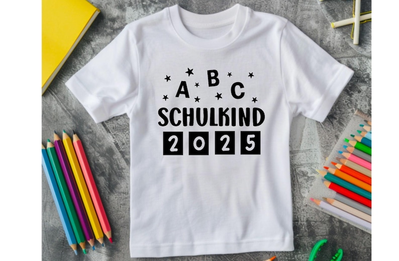 May include: White t-shirt with the text "A B C SCHULKIND 2025" in black. The shirt is surrounded by colored pencils, a yellow book, and a clear pencil case. The text is decorated with small black stars.