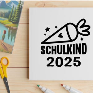 May include: A white notebook with the words "SCHULKIND 2025" and a black graphic of a school cone with stars. The image also includes a colorful landscape painting, yellow scissors, and markers on a wooden surface.