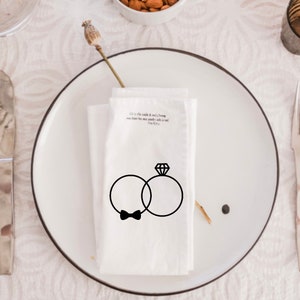 Op de afbeelding: Een witte servet met een zwarte illustratie van twee ringen en een strikje. De tekst "It is a little while & it will be long, you have been my steady rock & my song.  -The 1975" staat op de servet gedrukt.