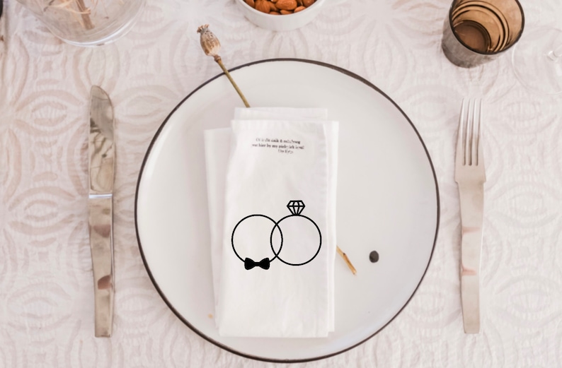 Op de afbeelding: Een witte servet met een zwarte illustratie van twee ringen en een strikje. De tekst "It is a little while & it will be long, you have been my steady rock & my song.  -The 1975" staat op de servet gedrukt.