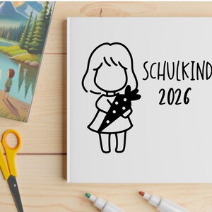 May include: A white notebook with a black illustration of a child holding a carrot, with the text "SCHULKIND 2026". The image also includes a colorful landscape painting, yellow scissors, and markers.