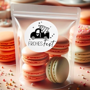 Puede incluir: Una bolsa de pl&aacute;stico transparente llena de macarons de color rosa pastel y verde. Una pegatina blanca con un cami&oacute;n negro que lleva un &aacute;rbol de Navidad y el texto "FROHES FEST" est&aacute; en la bolsa.