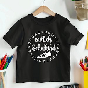 May include: Black short-sleeved t-shirt with a white circular design. The design includes the words "endlich Schulkind" and a carrot illustration. The shirt is hanging on a wooden hanger.