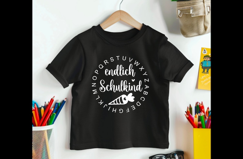 May include: Black short-sleeved t-shirt with a white circular design. The design includes the words "endlich Schulkind" and a carrot illustration. The shirt is hanging on a wooden hanger.