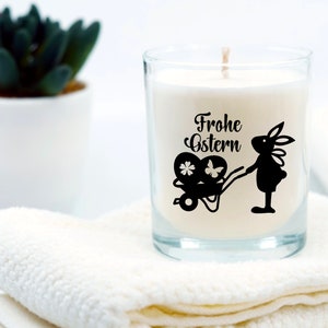 May include: A white candle in a clear glass jar with a black silhouette of a bunny pulling a wagon with Easter eggs. The candle has the text "Frohe Ostern" printed on it.