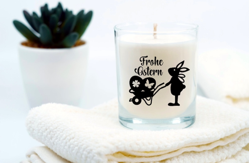 May include: A white candle in a clear glass jar with a black silhouette of a bunny pulling a wagon with Easter eggs. The candle has the text "Frohe Ostern" printed on it.