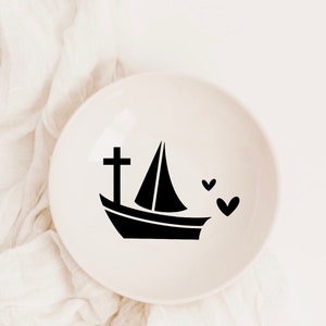 May include: A white ceramic dish with a black silhouette of a sailboat with a cross on the bow and three hearts.