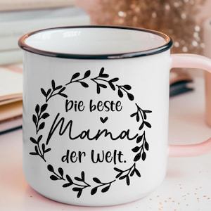 May include: White enamel mug with a black rim and pink handle. The mug has a black floral design with the text "Die beste Mama der Welt."