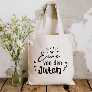 May include: A white tote bag with black text that reads "Eine von den Guten" which translates to "One of the good ones".