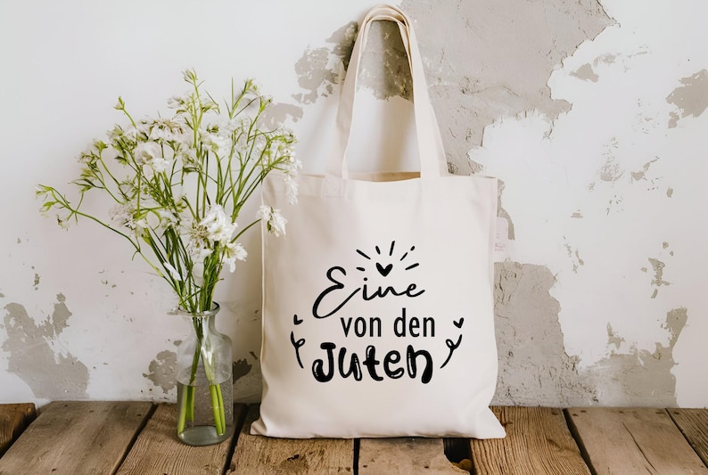 May include: A white tote bag with black text that reads "Eine von den Guten" which translates to "One of the good ones".
