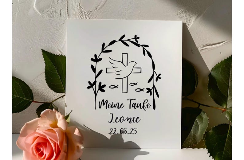 May include: A white square card with a black floral design, cross, dove, and fish. The text reads "Meine Taufe Leonie 22.05.25". A pink rose and green leaves are in the foreground.