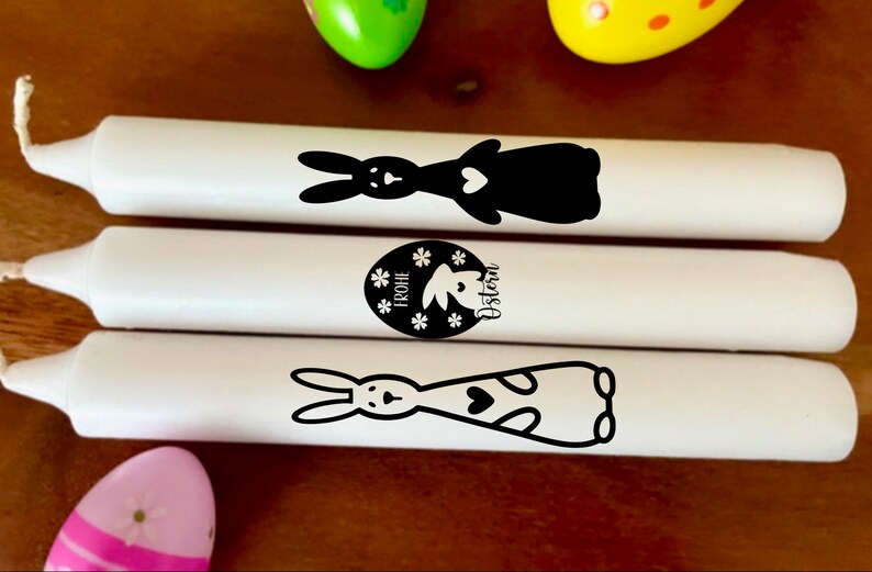 May include: Three white candles with black bunny and Easter egg designs. The candles are decorated with the text "Frohe Ostern" which means "Happy Easter" in German.