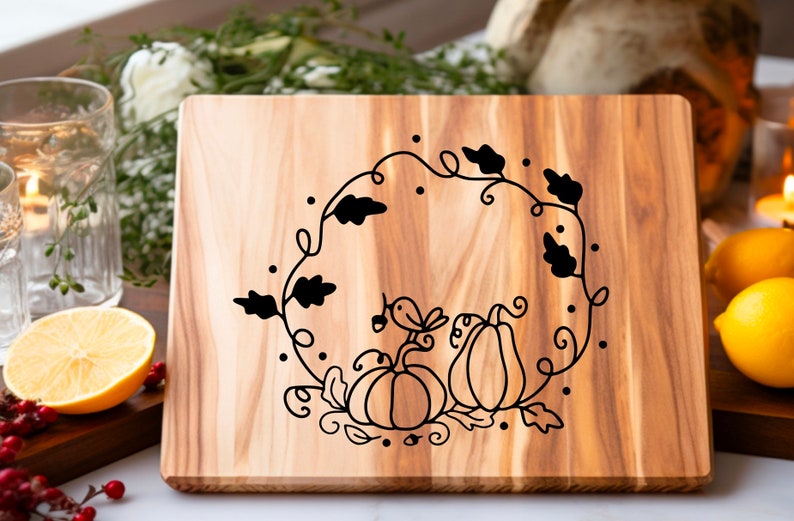 Op de afbeelding: Een houten snijplank met een zwart-wit ontwerp van een krans met pompoenen en een vogel. De krans is gemaakt van bladeren en wijnstokken.