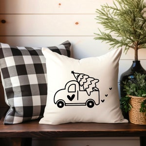 May include: A white throw pillow with a black and white illustration of a pickup truck carrying a Christmas tree. The truck has a heart on the side and stars on the wheels.
