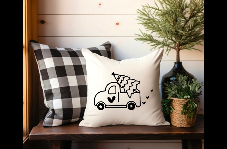 May include: A white throw pillow with a black and white illustration of a pickup truck carrying a Christmas tree. The truck has a heart on the side and stars on the wheels.