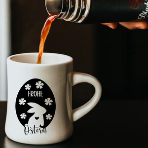 May include: A white ceramic mug with a black and white design of an Easter bunny inside an egg with the text "Frohe Ostern" on it. Coffee is being poured into the mug from a black thermos.