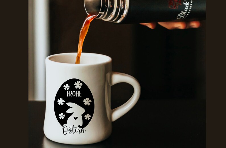 May include: A white ceramic mug with a black and white design of an Easter bunny inside an egg with the text "Frohe Ostern" on it. Coffee is being poured into the mug from a black thermos.