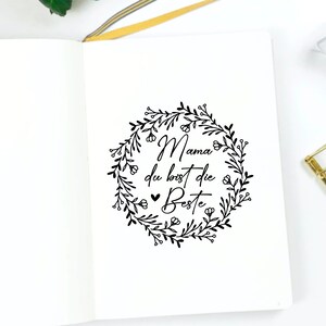 May include: A white notebook with a black floral wreath and the text "Mama du bist die Beste" inside the wreath.
