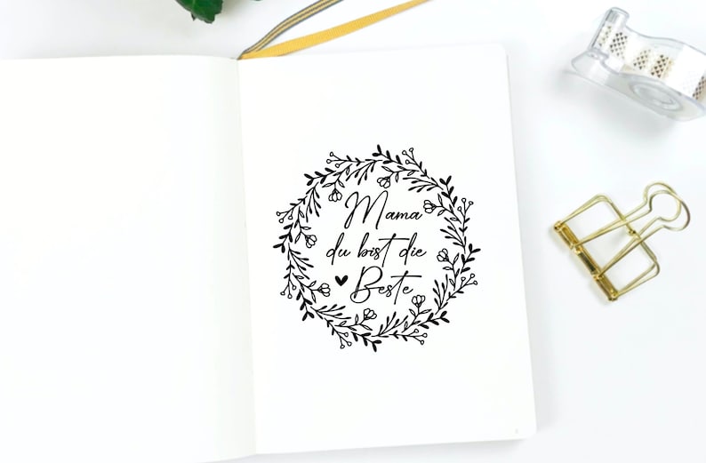 May include: A white notebook with a black floral wreath and the text "Mama du bist die Beste" inside the wreath.
