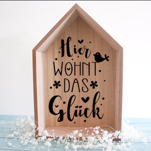 May include: A wooden house-shaped sign with the text "Hier wohnt das Gl&uuml;ck" (Here lives happiness) in black lettering. The sign also features a small bird silhouette and hearts.