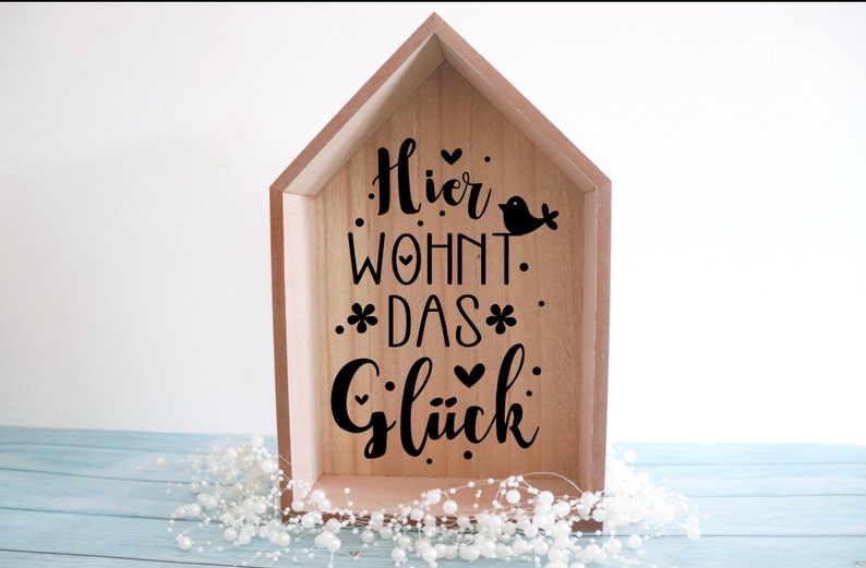 May include: A wooden house-shaped sign with the text "Hier wohnt das Gl&uuml;ck" (Here lives happiness) in black lettering. The sign also features a small bird silhouette and hearts.