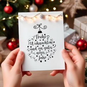 May include: A white card with black calligraphy that reads "Frohe Weihnachten und ein gutes neues Jahr"  The card is being held by a person's hands in front of a Christmas tree with red ornaments and lights.