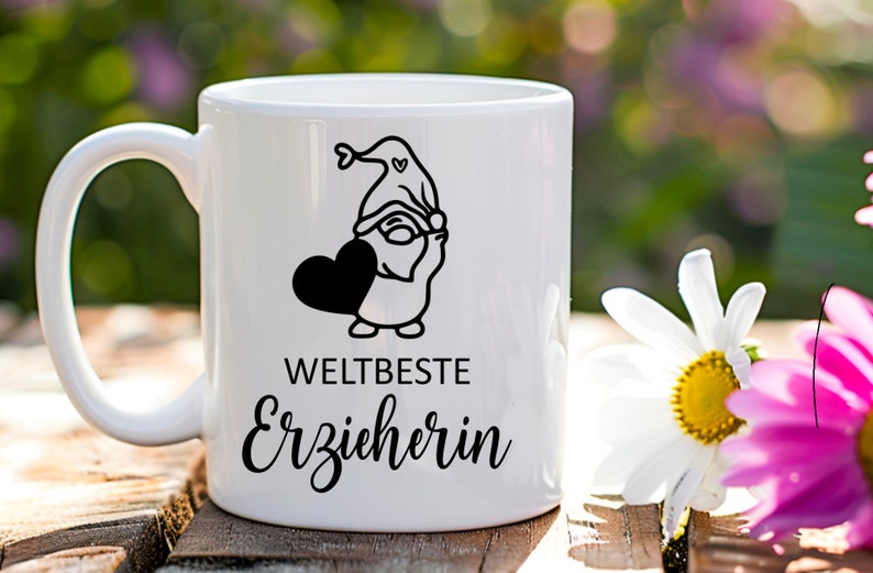 K&ouml;nnte beinhalten: Wei&szlig;e Keramiktasse mit einer schwarz-wei&szlig;en Illustration eines Zwergs, der ein Herz h&auml;lt. Der Text "WELTBESTE ERZIEHERIN" ist auf die Tasse gedruckt.