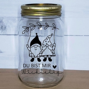 K&ouml;nnte beinhalten: Ein klares Glasgef&auml;&szlig; mit goldenem Deckel, das eine Schwarzwei&szlig;illustration von zwei Gnomen auf einer Schaukel zeigt. Das Gef&auml;&szlig; tr&auml;gt den Text "Du bist mir wichtig" in schwarzer Schrift.