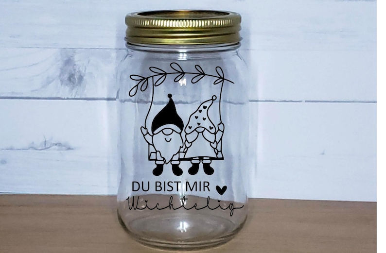 K&ouml;nnte beinhalten: Ein klares Glasgef&auml;&szlig; mit goldenem Deckel, das eine Schwarzwei&szlig;illustration von zwei Gnomen auf einer Schaukel zeigt. Das Gef&auml;&szlig; tr&auml;gt den Text "Du bist mir wichtig" in schwarzer Schrift.