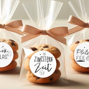 May include: Three clear cellophane bags tied with brown ribbon, each containing a stack of chocolate chip cookies with a round white sticker on top. The stickers have black text and illustrations. The stickers read "Gl&uuml;ck im Glas", "Zimtstern Zeit", and "Pl&auml;tzchen Fee".