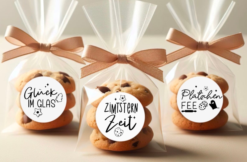 May include: Three clear cellophane bags tied with brown ribbon, each containing a stack of chocolate chip cookies with a round white sticker on top. The stickers have black text and illustrations. The stickers read "Gl&uuml;ck im Glas", "Zimtstern Zeit", and "Pl&auml;tzchen Fee".