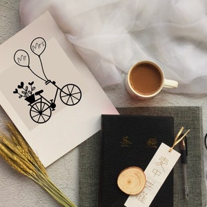 Op de afbeelding: Een witte kaart met een zwart-witte illustratie van een fiets met een mand vol bloemen en twee ballonnen met daarop "Mr" en "Mrs". De kaart ligt op een grijs oppervlak met een kopje koffie, een boek, een bril en tarwehalmen.