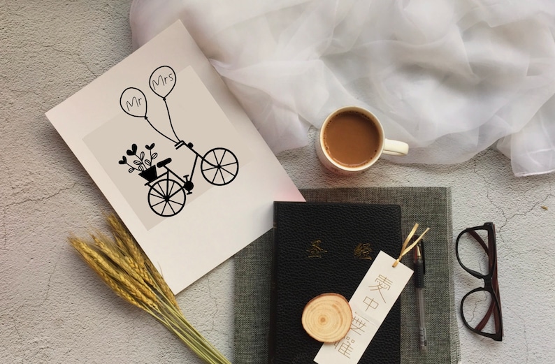 Op de afbeelding: Een witte kaart met een zwart-witte illustratie van een fiets met een mand vol bloemen en twee ballonnen met daarop "Mr" en "Mrs". De kaart ligt op een grijs oppervlak met een kopje koffie, een boek, een bril en tarwehalmen.