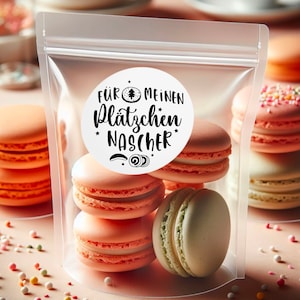 May include: A clear plastic bag filled with six pastel-coloured macarons. A white round sticker with black text reads "F&Uuml;R MEINEN PL&Auml;TZCHEN NASCHER" with a small Christmas tree icon.