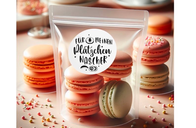 May include: A clear plastic bag filled with six pastel-coloured macarons. A white round sticker with black text reads "F&Uuml;R MEINEN PL&Auml;TZCHEN NASCHER" with a small Christmas tree icon.
