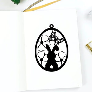May include: Black silhouette of a bunny rabbit inside an egg shape with flowers and a butterfly. The design is perfect for a spring or Easter craft project.