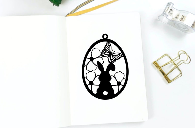 May include: Black silhouette of a bunny rabbit inside an egg shape with flowers and a butterfly. The design is perfect for a spring or Easter craft project.