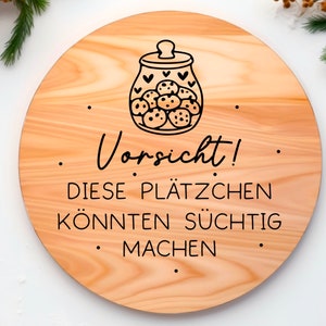 May include: A round wooden sign with a black and white illustration of a cookie jar filled with cookies. The sign reads "Vorsicht! Diese Pl&auml;tzchen k&ouml;nnten s&uuml;chtig machen."
