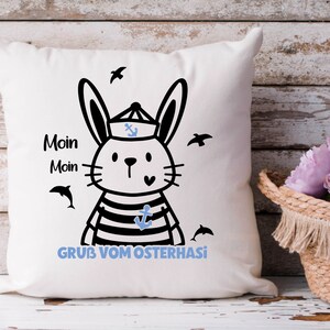 May include: White pillow with a black and white illustration of a bunny wearing a sailor hat and striped shirt. The bunny is holding a blue anchor. The text "Moin Moin GRUB VOM OSTERHASI" is printed on the pillow.