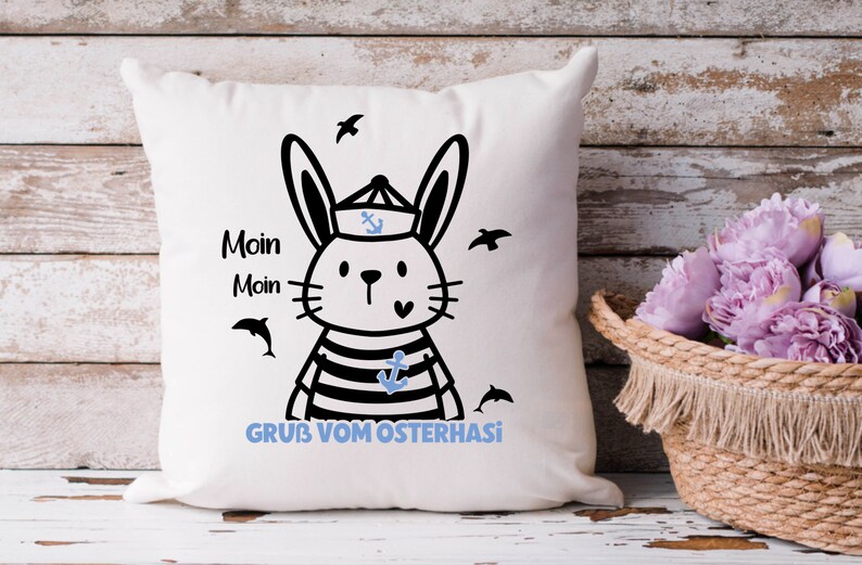 May include: White pillow with a black and white illustration of a bunny wearing a sailor hat and striped shirt. The bunny is holding a blue anchor. The text "Moin Moin GRUB VOM OSTERHASI" is printed on the pillow.