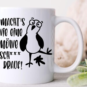 May include: White ceramic mug with a black cartoon seagull and German text. The text reads "MACHT'S WIE EINE M&Ouml;WE SCH*** DRAUF!" The seagull has large eyes and is in a comical pose. The mug has a curved handle.