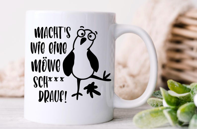 May include: White ceramic mug with a black cartoon seagull and German text. The text reads "MACHT'S WIE EINE M&Ouml;WE SCH*** DRAUF!" The seagull has large eyes and is in a comical pose. The mug has a curved handle.
