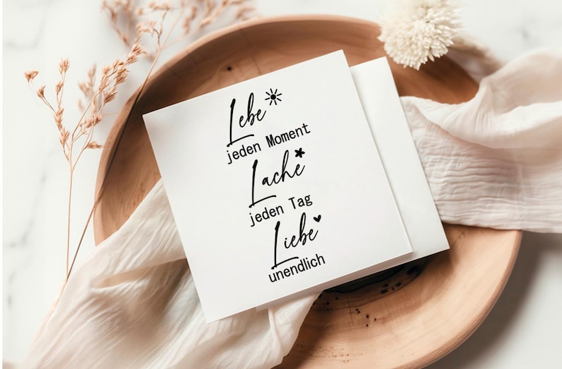 May include: White card with black text that reads "Liebe jeden Moment Lache jeden Tag Liebe unendlich". The card is on a wooden surface with a white cloth.