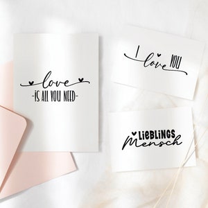 Puede incluir: Tres tarjetas blancas con caligraf&iacute;a negra. Las tarjetas dicen "love is all you need", "I love you" y "Lieblings Mensch".
