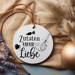 May include: White round ornament with black text and illustrations. The text reads "Zutaten voller Liebe" which translates to "Ingredients full of love". The illustrations include a whisk, a heart, and a candy.