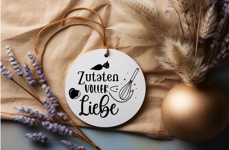 May include: White round ornament with black text and illustrations. The text reads "Zutaten voller Liebe" which translates to "Ingredients full of love". The illustrations include a whisk, a heart, and a candy.