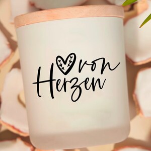May include: White candle with a wooden lid and a black heart with leaves design. The text "von Herzen" is written in black script on the candle.