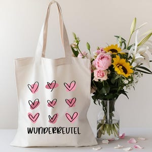 May include: White canvas tote bag with pink and black hearts and the text "WUNDERBEUTEL". The bag is next to a vase of flowers.