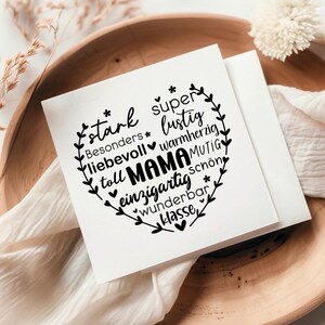 May include: A white card with a black heart-shaped design featuring the word "Mama" surrounded by positive adjectives in German. The card is on a wooden surface with a white fabric and a white flower.